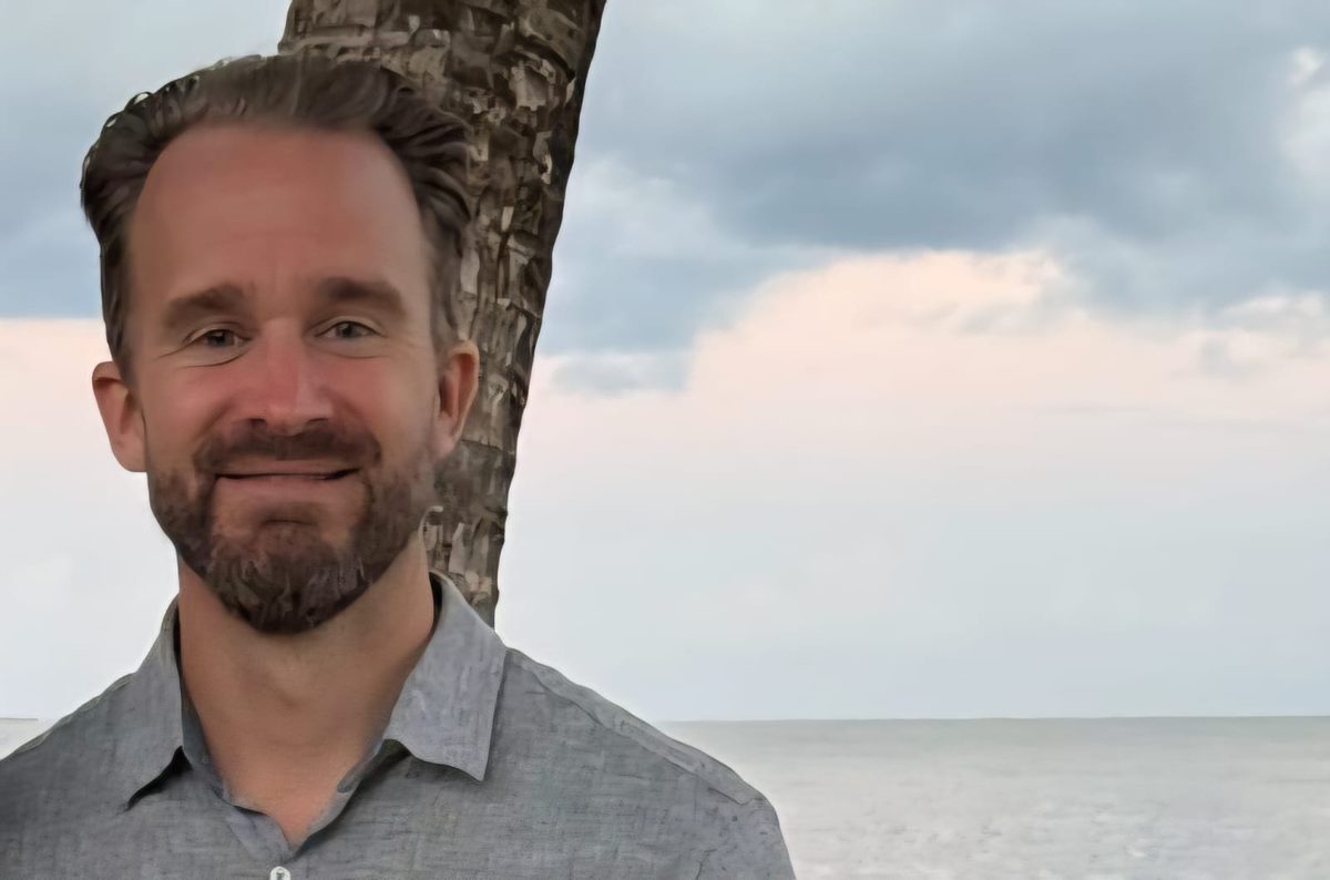 Simo D smiling on a beach in Belize, with a palm tree and the Caribbean Sea behind him